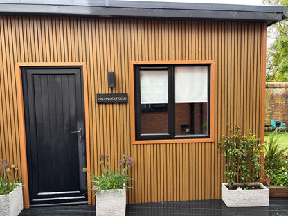 A garden room clad in composite cedar slat cladding with composite black door, matching black composite decking and light grey planters newly planted.