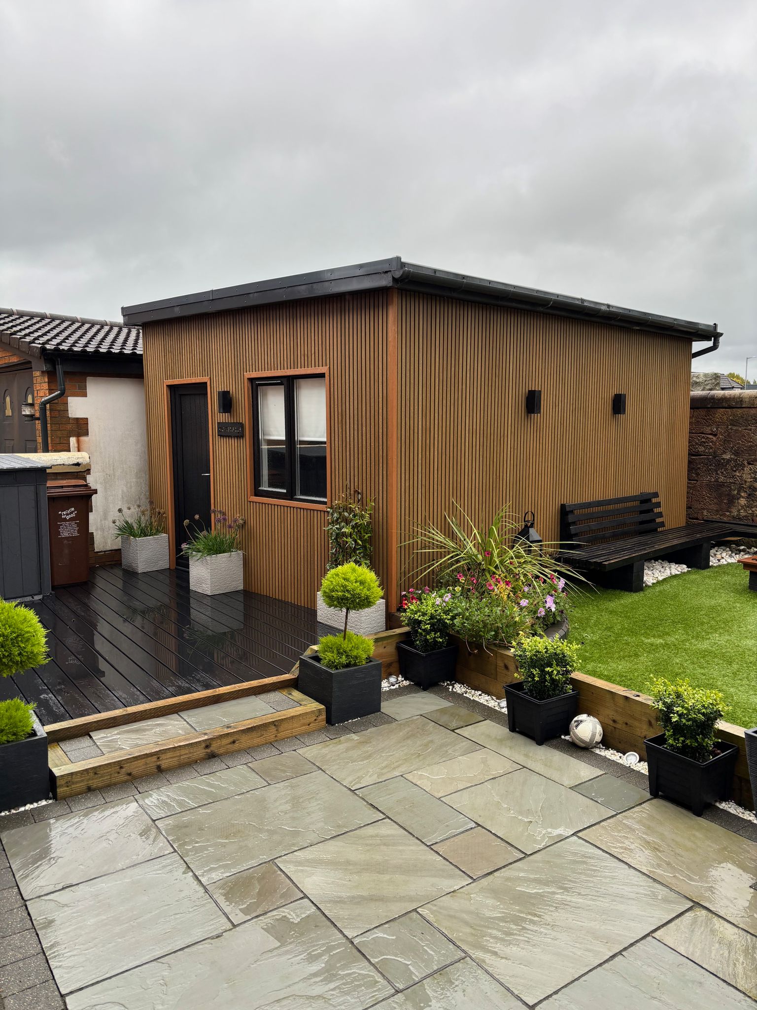 A Cedar composite slat clad garden building sat in a newly landscaped garden with black composite decking, artificial lawn area and grey porcelain paving all furnished with seating area and a selection of planters.