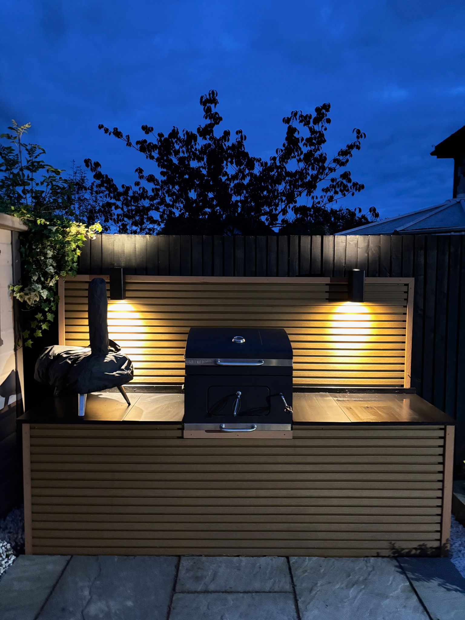 A lovely BBQ station set up in the garden patio area clad in Cedar composite slatted cladding with black warm white down lighters and a pizza oven and grill.