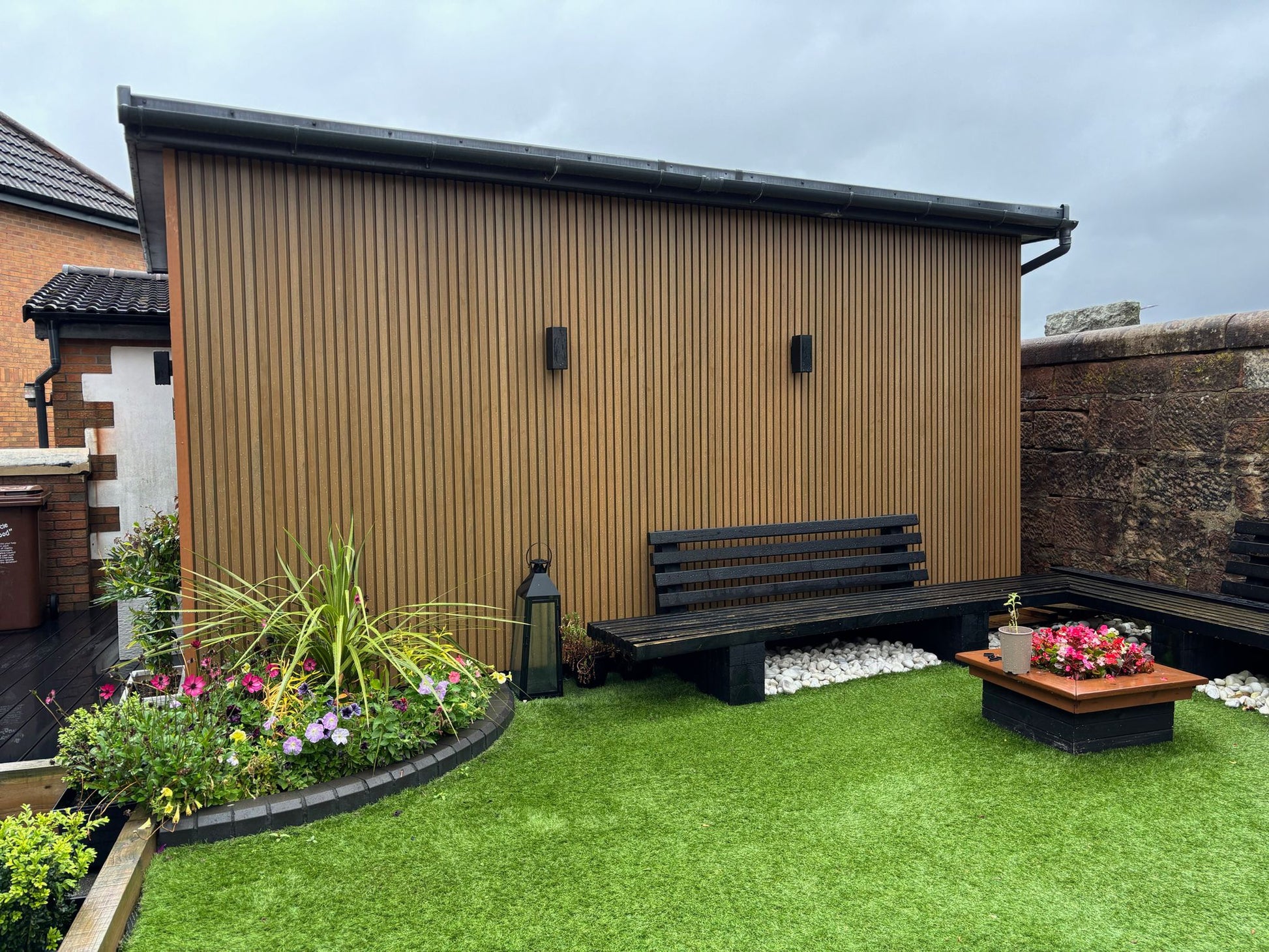 The side elevation of a garden room clad with Cedar composite slat cladding, showing the seating area with freshly planted flower beds and artificial grass.