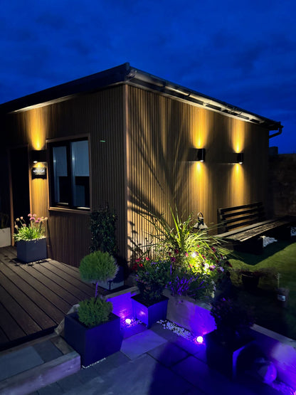 An evening picture of a garden room clad in Cedar sometimes known as spiced oak composite slat cladding, with warm white down lighters in a newly landscaped garden.