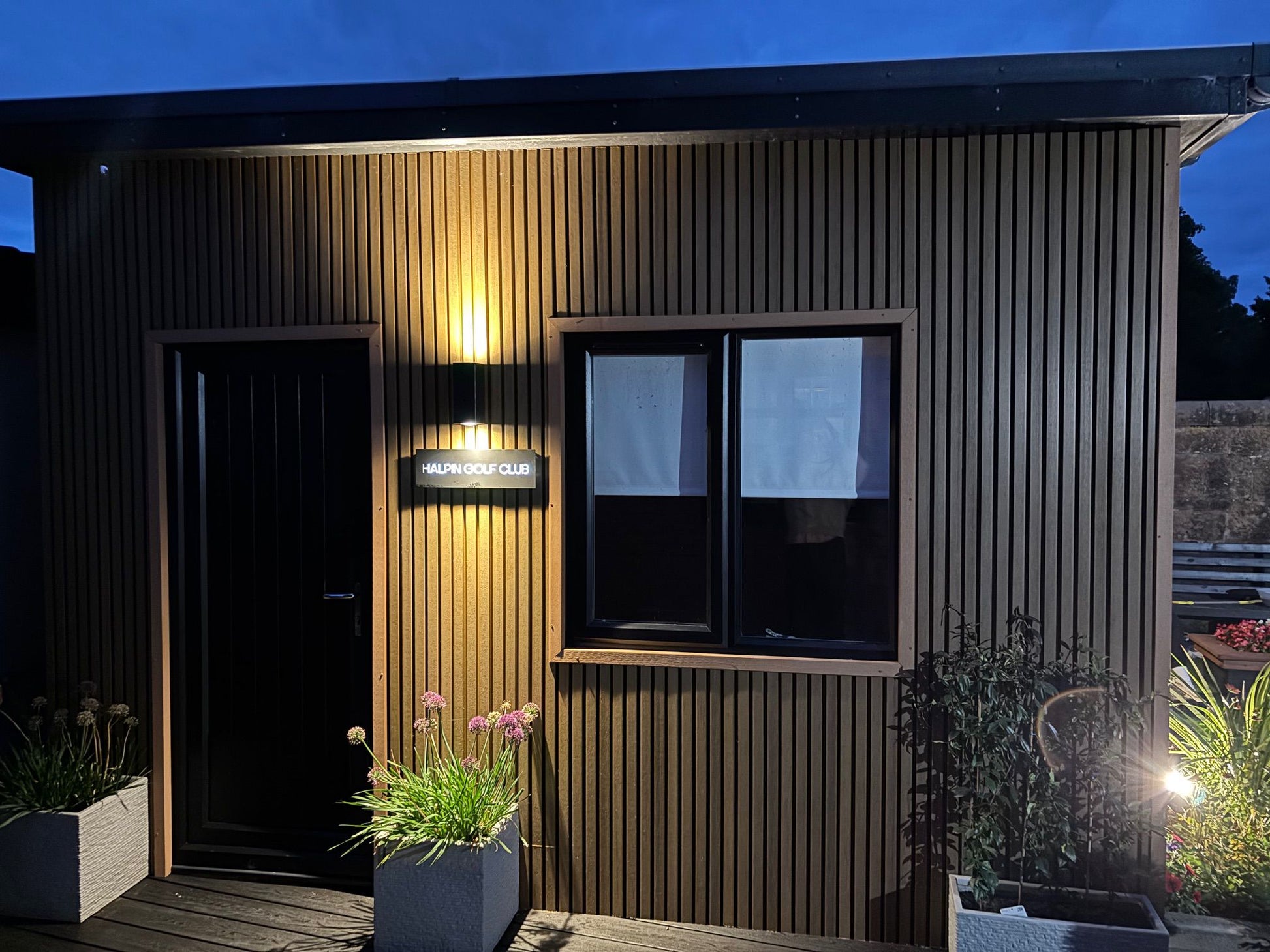 An evening picture of our customers garden room clad in Cedar composite slat cladding with warm white down lighters and charcoal black, composite decking.