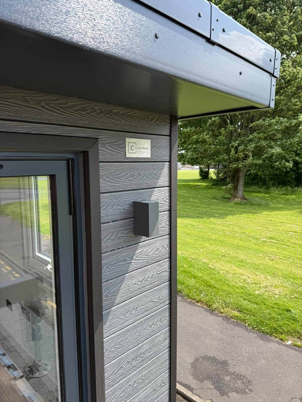 Modern building exterior with glass door and wooden paneling, green grass and trees in the background.