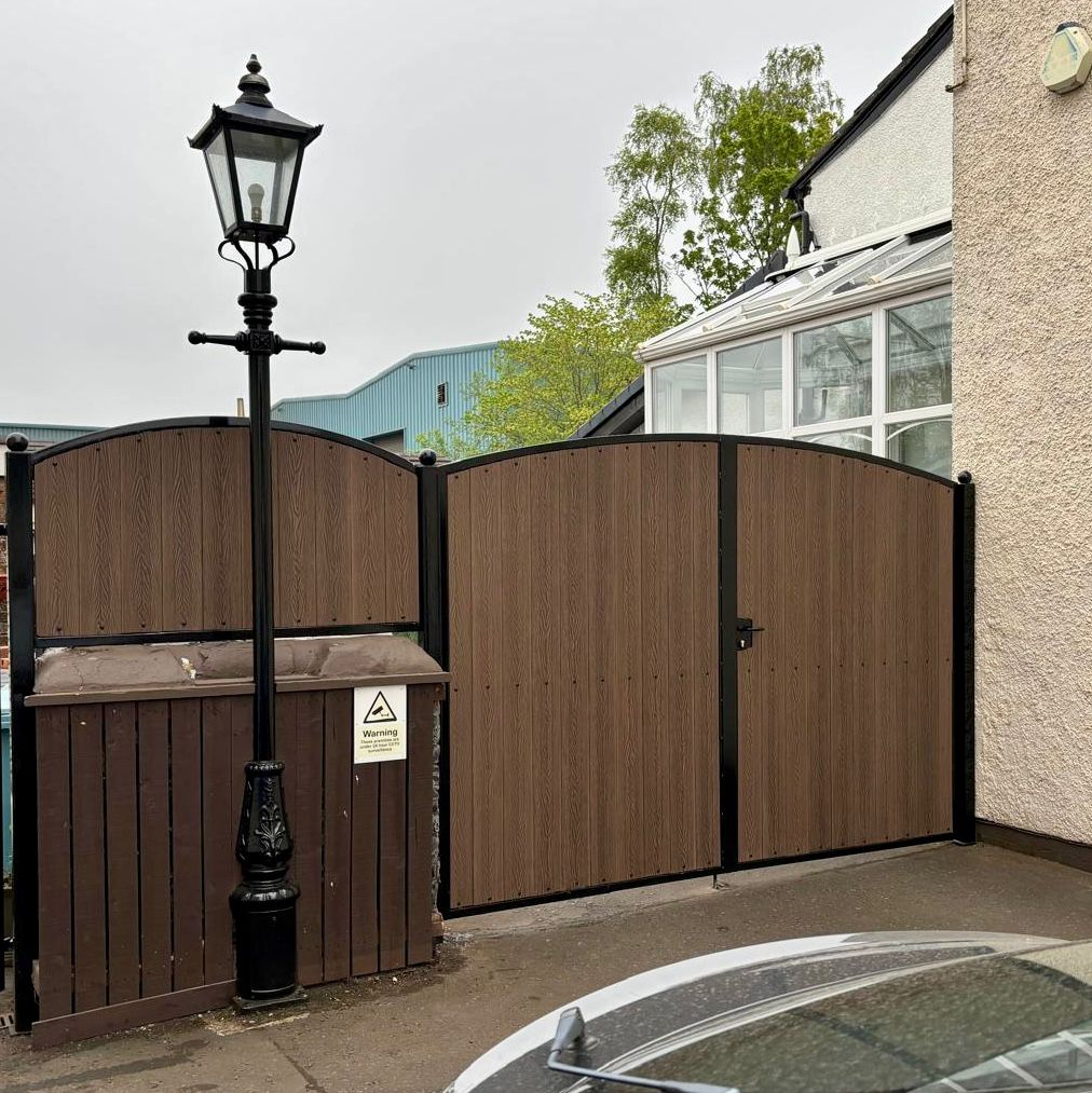 A 6ft high set of dark oak composite driveway gates with black powder coated & galvanised steel frame, with a matching side fence panel and a Victorian style lampost. 