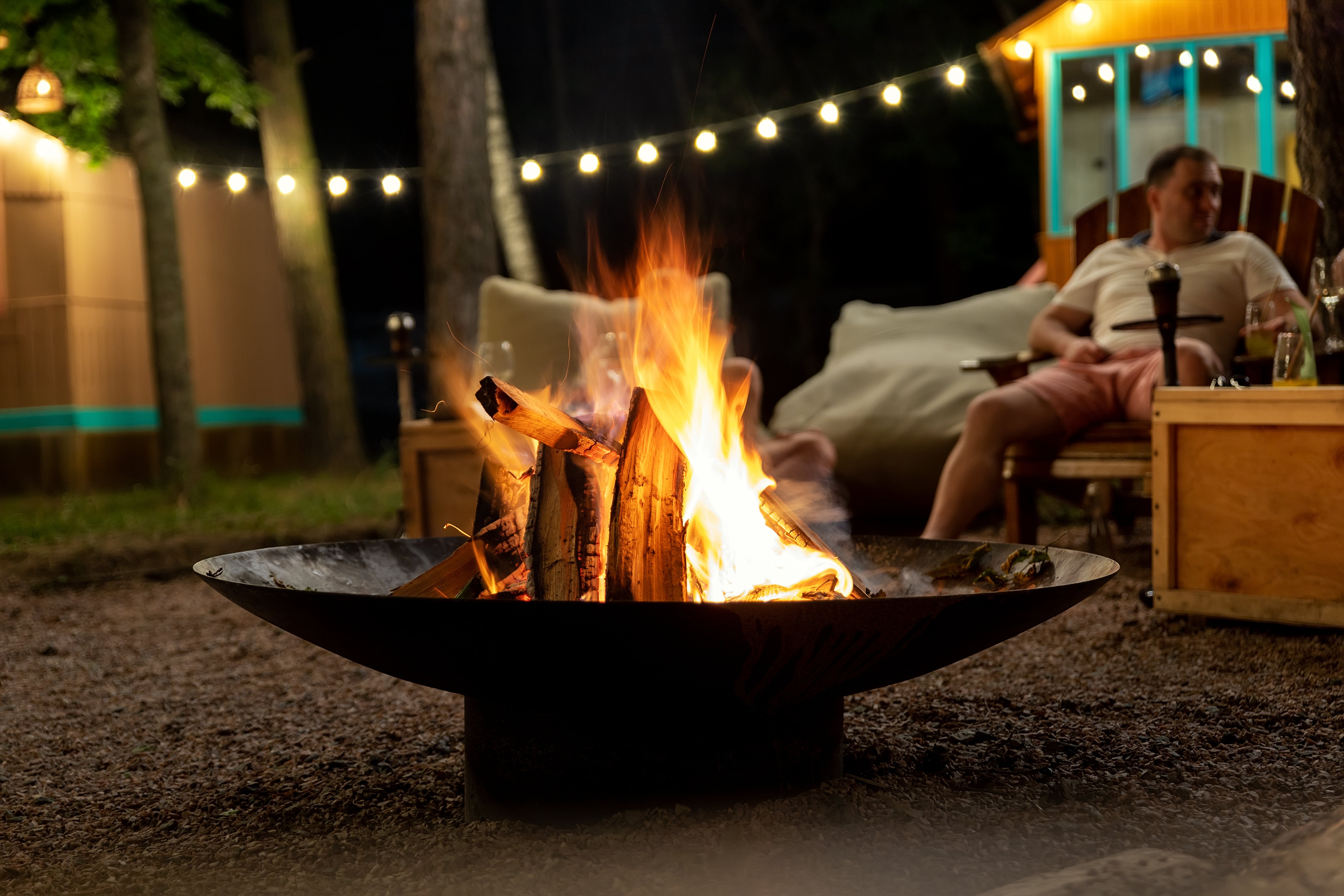 A man sits outside socialising with friends in front of a fire pit heaped burning with logs.
