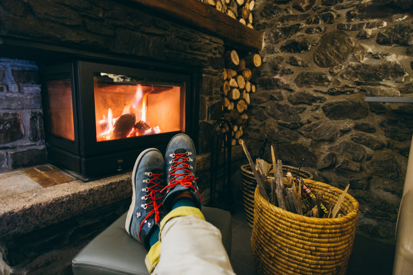 Someone sitting with there feet up in front of a log burning stove, with log basket next to them.