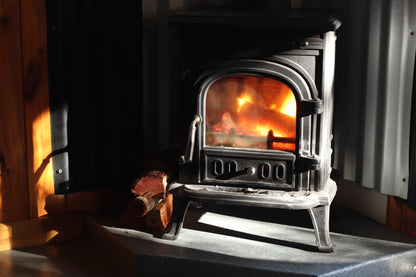 A traditional firewood stove burning away in a dark room cast in light from a window.