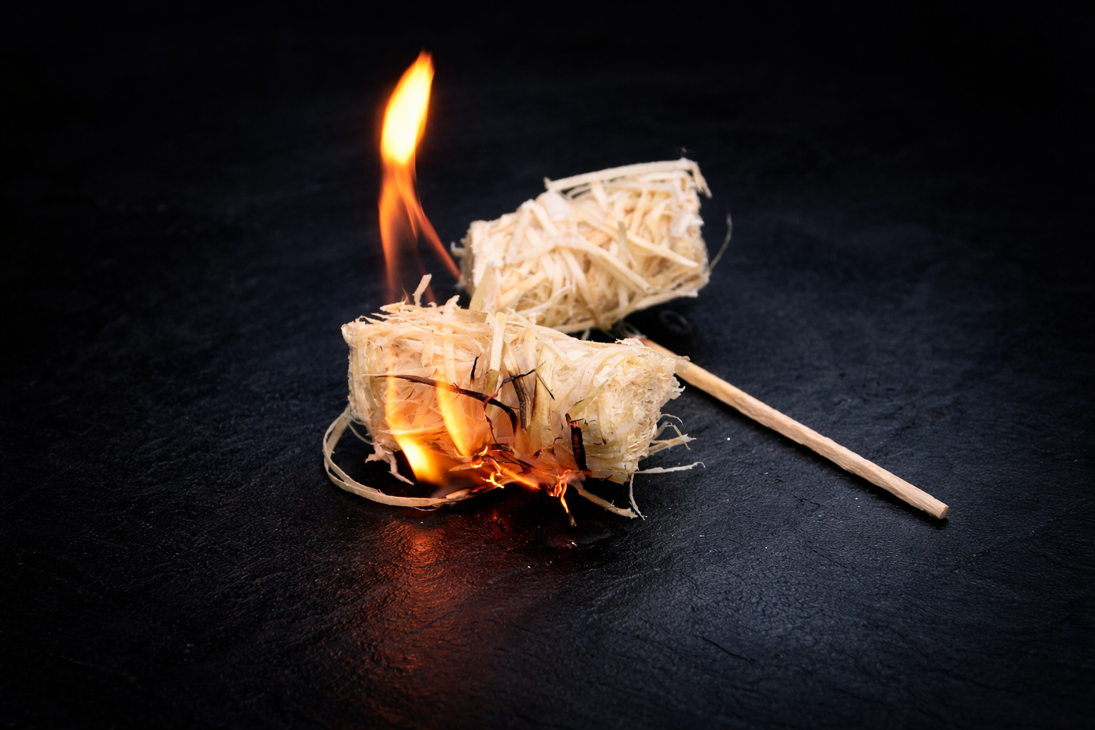 Two firelighters on a dark surface with one lit, emitting flames.