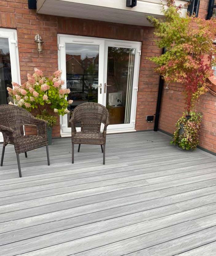 A modern composite decking installation with grey wood-like appearance, featuring two wicker chairs on top, flanked by green and red plants, with a house and large windows in the background.